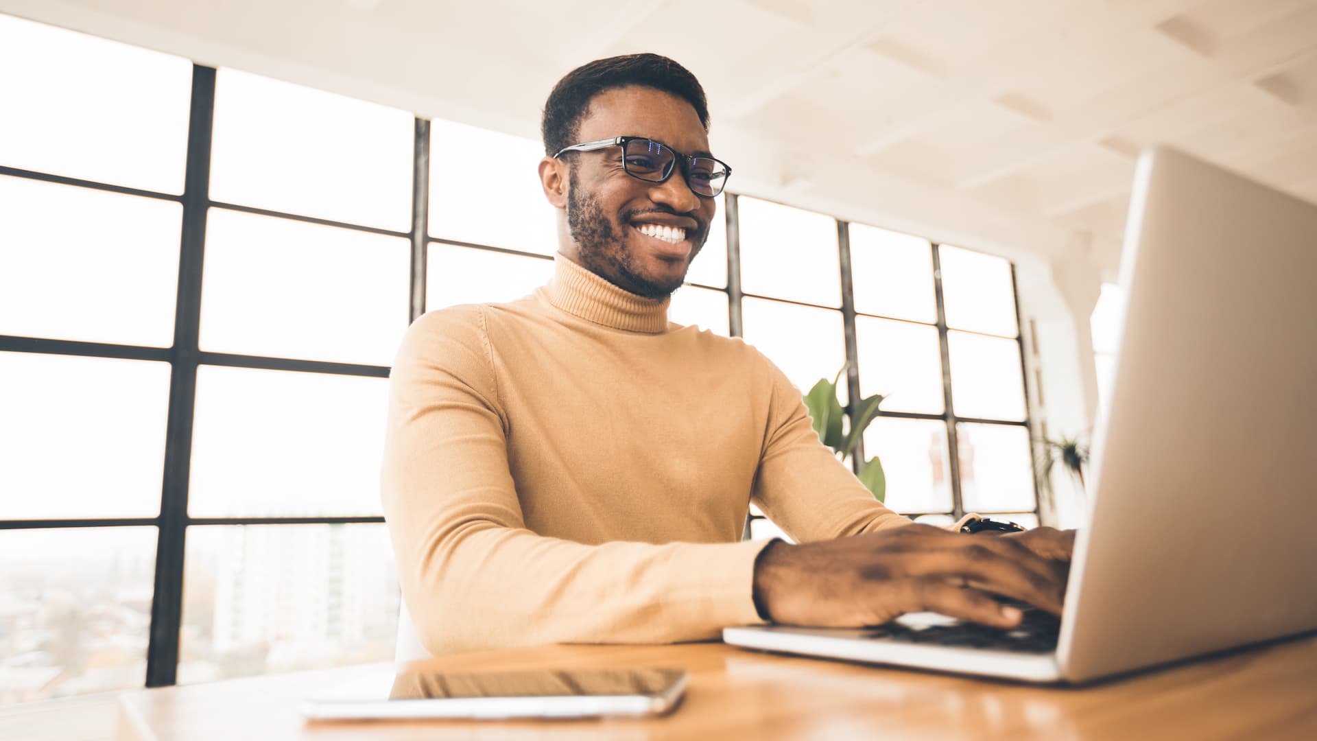 Smiling man wearing a sweater while looking at laptop on wood desk. 